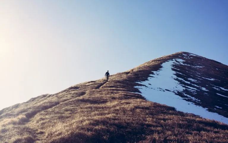 Ein männlicher Wanderer, der allein einen steilen Bergpfad im Winter hinabsteigt. Aus der Ferne aufgenommen. Matte hohe Kontrastwirkung
