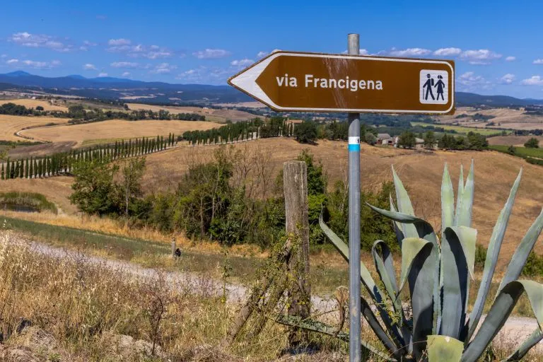 Landschaft entlang der Via Francigena mit Schlammstraße, Feldern, Bäumen und Weinberg.  Schild, das die Richtung von Monteroni d'Arbia anzeigt, Route der Via Francigena. Provinz Siena, Toskana. Italien, Europa.